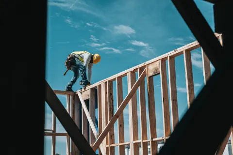 Construction worker wearing a safety helmet working on framing a wooden house under a clear blue sky.