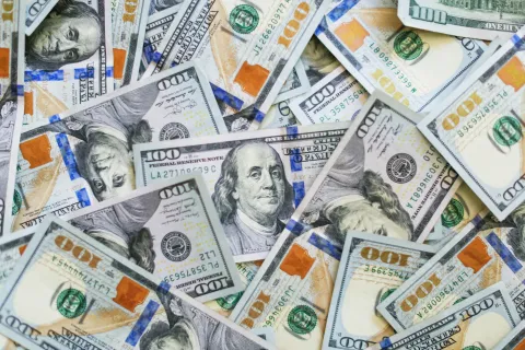 Pile of US one hundred dollar bills overlapping, showing detailed security features and Benjamin Franklin portrait.