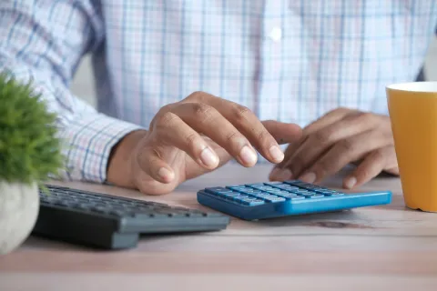 A person calculating with a blue calculator beside a yellow cup and potted plant on a wooden desk.