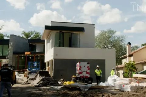 Modern two-story house under construction with workers, construction materials, and clear sky in a residential area.
