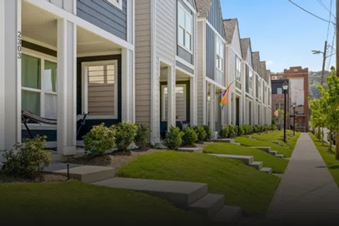 Row of modern townhouses with porches, manicured lawns, and a sidewalk under a clear blue sky.