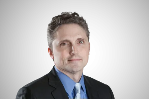 Professional middle-aged man with gray hair wearing a dark suit, blue shirt, and patterned tie on black background