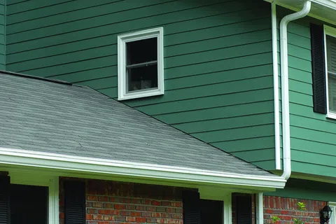 Green siding walls with white trim and a small window above a grey asphalt shingle roof on a residential house.
