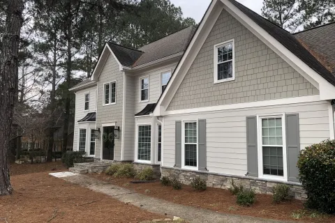 Large two-story suburban house with gray siding, white trim, stone foundation, and surrounding pine trees.