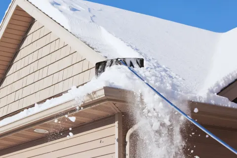 Person using a long snow shovel to clear heavy snow off a beige house roof on a clear winter day.