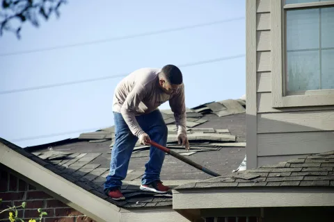 Man removing old shingles from a roof with a roofing shovel on a sunny day.