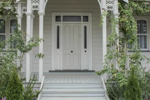 White wooden Victorian house entrance with detailed porch, stairs, and surrounding greenery and flowers.