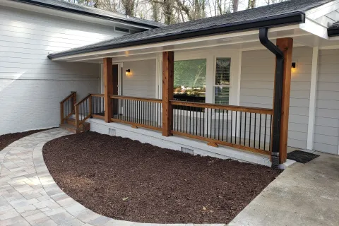 Modern porch with wooden railing, black metal balusters, and white exterior walls surrounded by mulch and paver walkway.