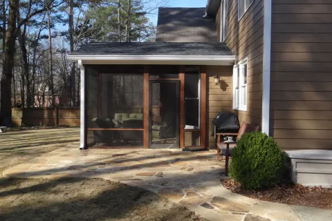 Stone patio with screened porch, outdoor grill, bench, and brown siding house surrounded by trees and yard.