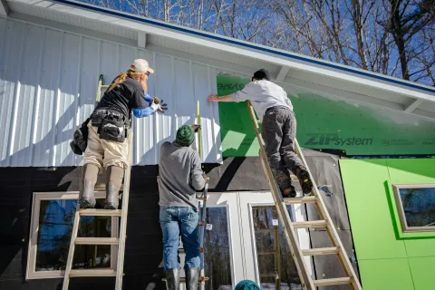 Three construction workers installing white siding on a house exterior under a clear blue sky.