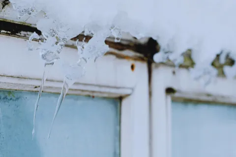 Icicles forming on a snow-covered, weathered window frame with frosted glass panes in cold weather.