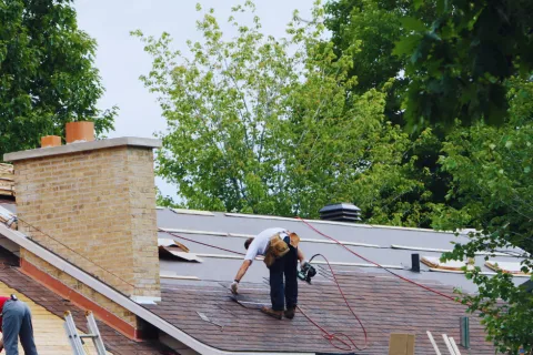 Roofers replacing shingles on a residential house roof surrounded by green trees on a clear day
