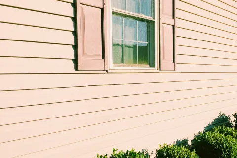 Beige house exterior with a window and shutters lined by neatly trimmed green bushes in bright sunlight.