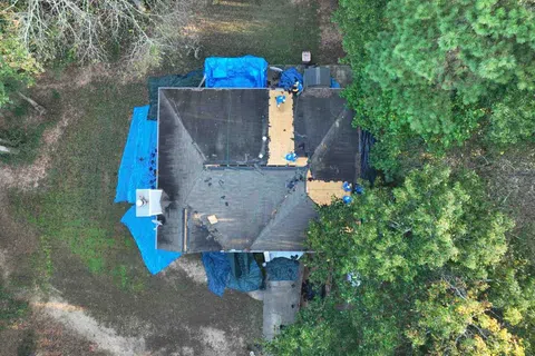 Aerial view of a house roof under repair with tarps and construction workers amidst surrounding trees.