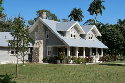 Two-story gray house with stone chimney, front porch, lush lawn, palm trees, and surrounding greenery on a sunny day