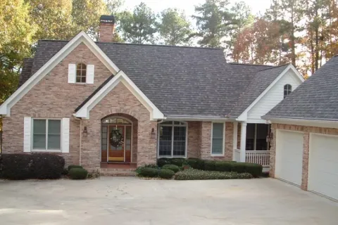 Brick house with gray shingled roof, white trim, front porch, garage, and surrounding greenery under daylight.
