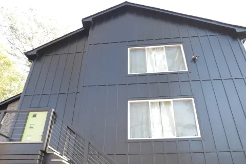 Modern dark gray house exterior with two large windows and a balcony with metal railing under bright daylight.