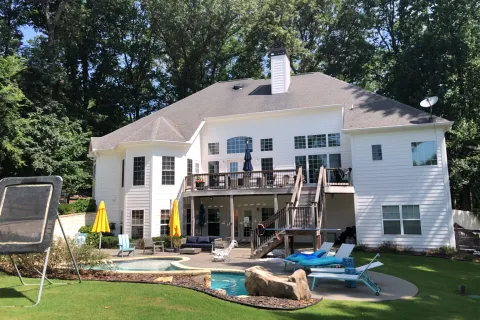 Spacious modern home with a pool, deck, and green backyard, surrounded by trees and sunny skies.