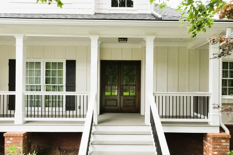 Front porch of a white house with wooden double doors, black railings, and brick foundation.
