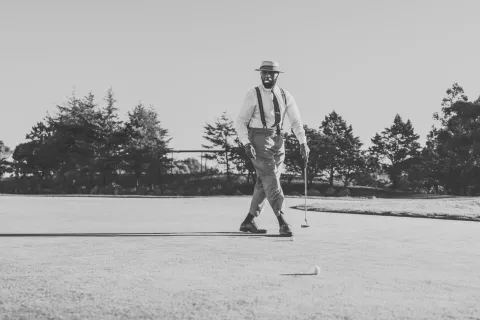 Black and white image of a man in vintage attire playing golf on a sunny course with trees in the background.