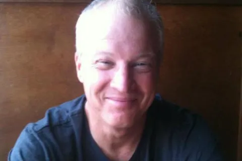 Smiling middle-aged man with short gray hair wearing a black t-shirt and silver watch sitting at a wooden table.