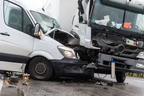 White van and truck with severe front-end damage after collision on wet road with debris scattered.