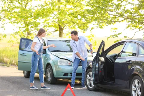 Two people arguing next to their cars after a minor collision on a sunny road with a warning triangle placed on the ground.