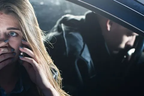 Distressed woman with a bruised face makes a phone call from a car while a man sits in the driver's seat.