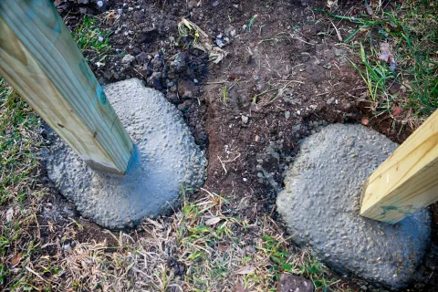 Close-up of wooden posts set in wet concrete, surrounded by soil and grass, showcasing construction work.