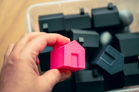 Hand holding a bright pink toy house above multiple black toy houses in a container on wooden surface.