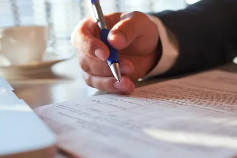 A close-up of a person signing paperwork at a desk, with a cup of coffee in the background.