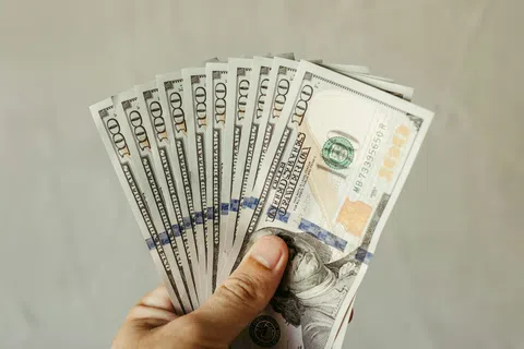 Hand holding a fan of multiple US hundred-dollar bills with a plain blurred background.