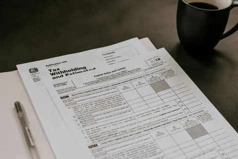 Tax withholding and estimated tax forms with pen on folder and coffee cup on dark surface.