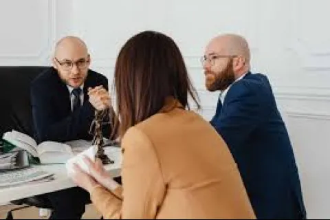 Two men in suits consulting a woman in a brown jacket at an office desk with legal books and scales of justice.