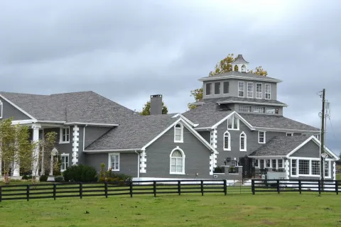 Large gray country estate with multiple peaked roofs surrounded by black fence and green lawn under cloudy sky