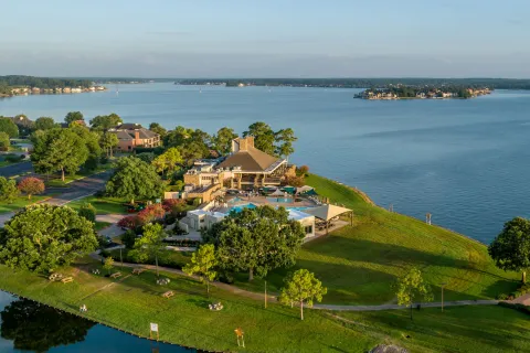 Aerial view of a lakeside resort with a pool, green lawns, trees, and nearby islands under a clear sky.