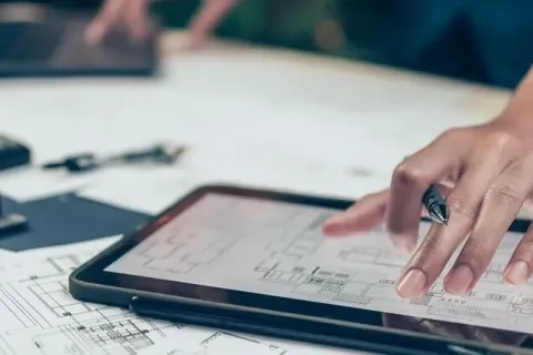 A hand pointing at a tablet displaying architectural plans on a workspace with tools.