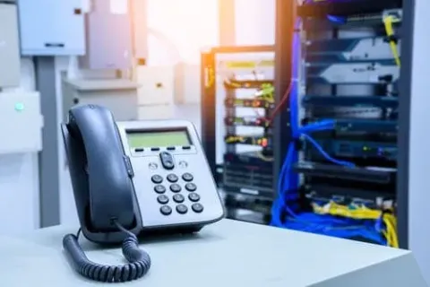 A telephone on a desk in a server room with networking equipment in the background.