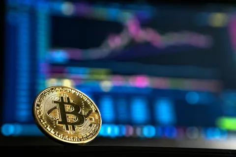 Close-up of a gold Bitcoin in front of colorful cryptocurrency charts on a screen.