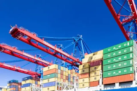 Colorful shipping containers stacked at a port with cranes against a clear blue sky.