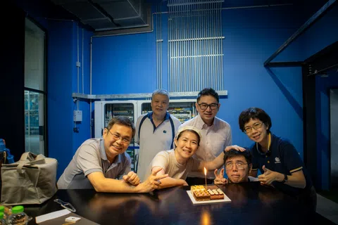 Group of six coworkers celebrating with a small birthday cake and candle in a modern office with blue walls.
