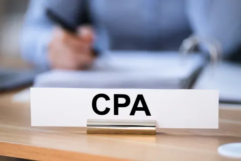 Nameplate with CPA text on a desk with a person writing blurred in the background