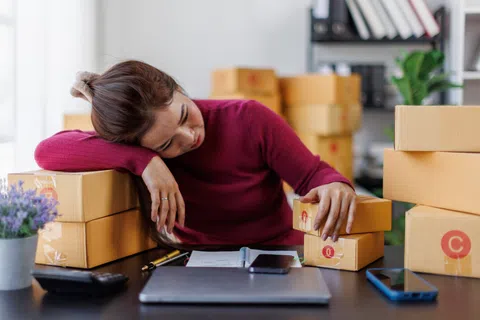 Tired woman resting head on arm amid stacked cardboard boxes and open notebook while working at desk.