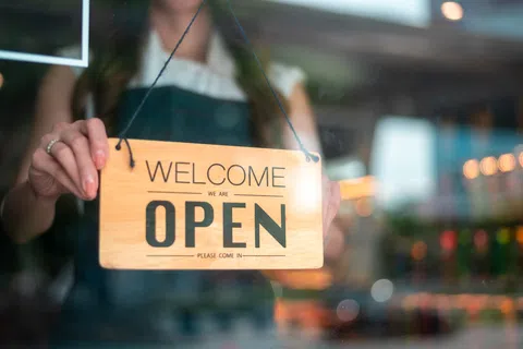 Person holding a wooden welcome sign that says open, inviting customers to enter a store or business.