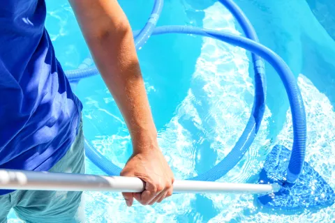 Person cleaning clear blue swimming pool water with a manual pool vacuum and hose on a sunny day
