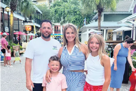 Smiling family of four posing under red and white umbrellas on a sunny shopping street with palm trees.
