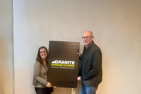 Two people holding a Granite Garage Floors sign in an indoor setting with textured floor and plain wall.