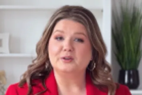 Woman in red blazer speaking in a modern office with white shelves and decorative plants in background.