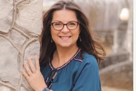 Smiling woman with glasses wearing a blue blouse and smartwatch leaning on stone wall outdoors