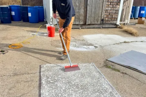 Man using a long-handled tool to spread material on a concrete surface outside near barrels and a wooden shed.